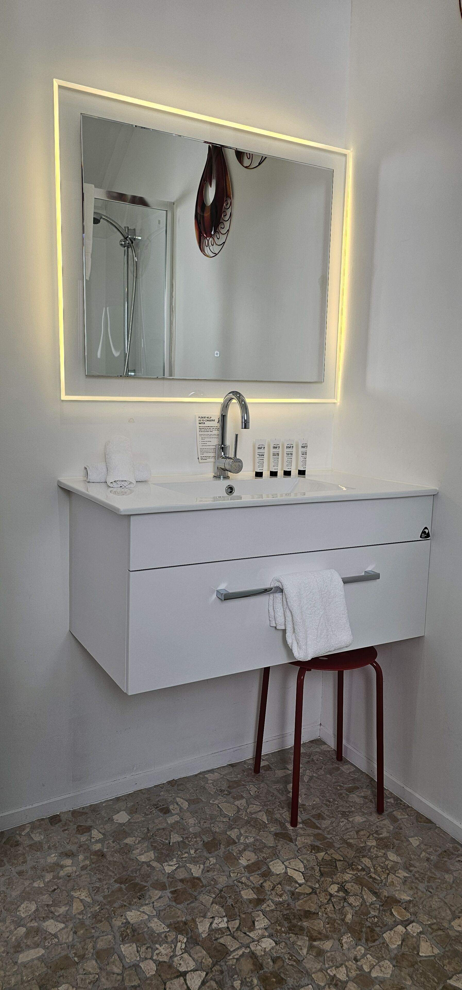Un baño moderno con tocador blanco, un espejo grande con iluminación indirecta y una ducha, además de un taburete pequeño.