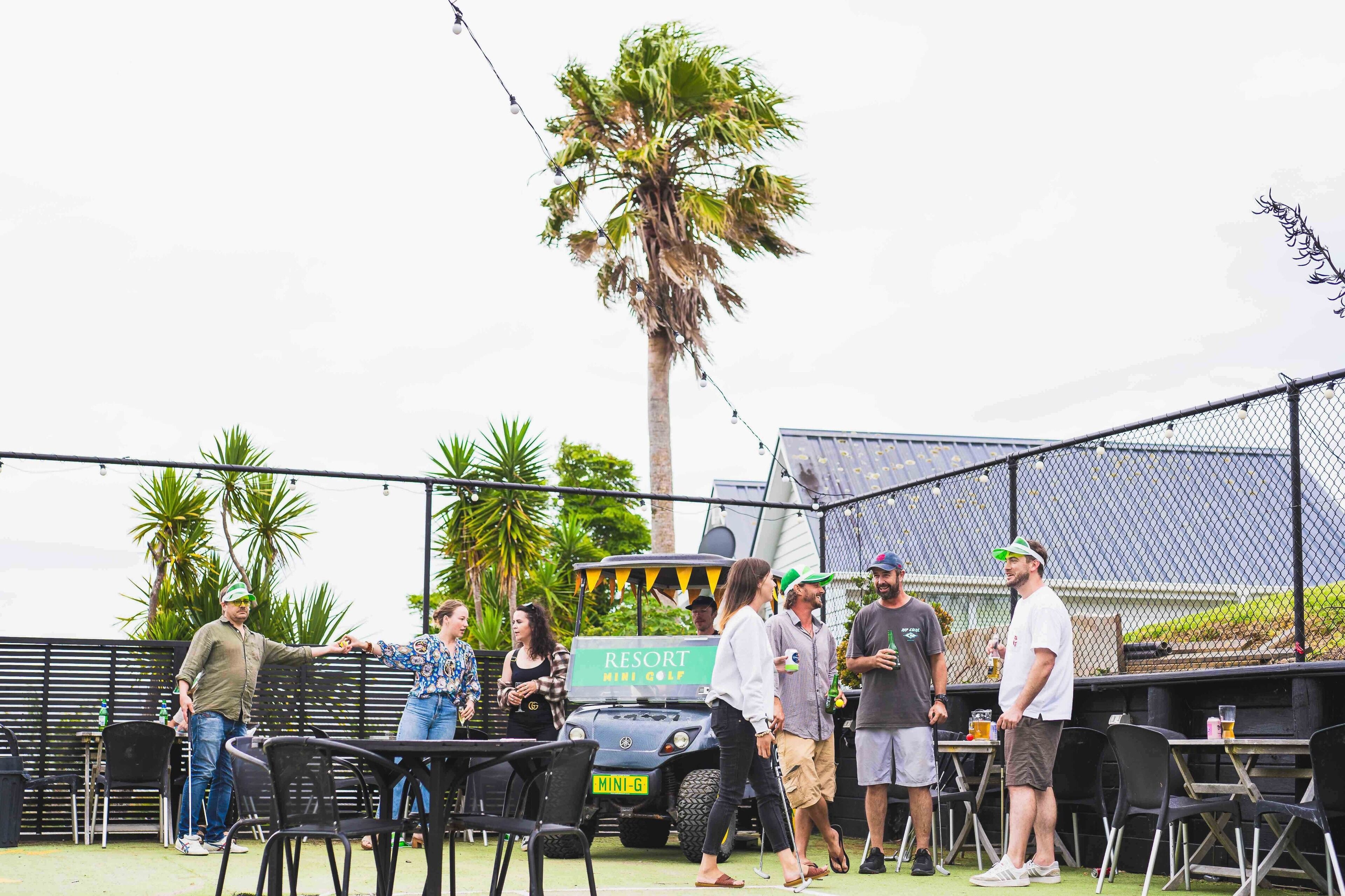 A group of people gathered around a golf cart labeled "RESORT MINI GOLF" in an outdoor setting with tables and chairs, palm trees, and a fence.