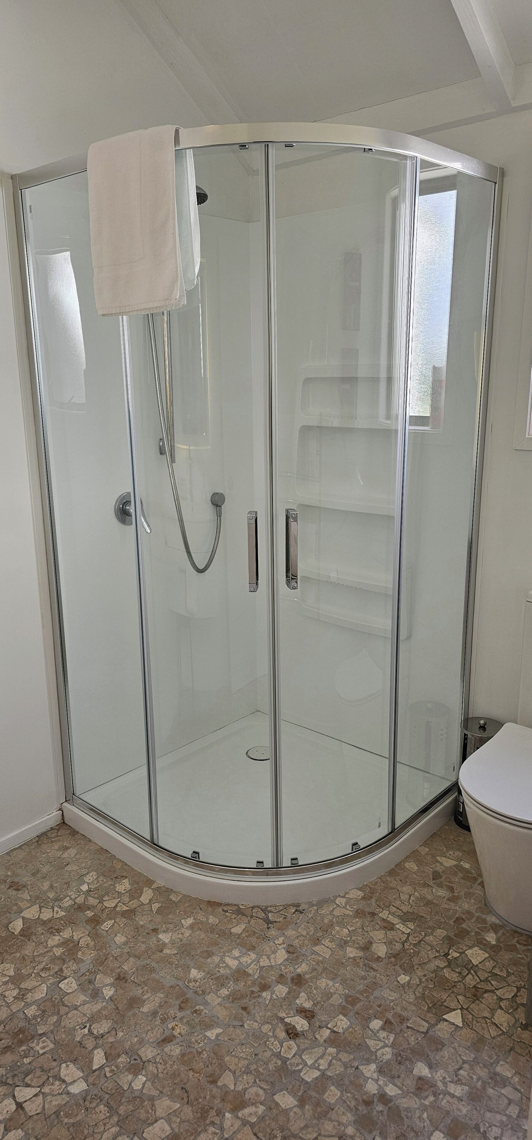 Ein Duschbereich mit Glaswänden, ein weißes Handtuch hängt an der Tür, ein gefliester Boden und eine Toilette auf der rechten Seite.