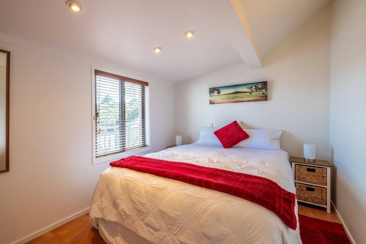 A bedroom with a bed, a window with blinds, a painting on the wall, and a bedside table with a lamp.