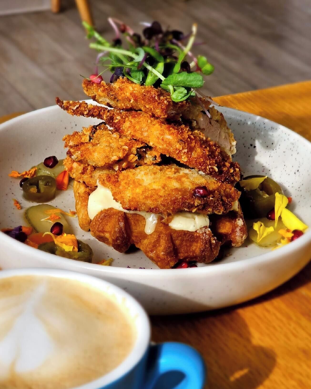 Une assiette d’ailes de poulet panées, nappées d’une sauce crémeuse, garnies de jeunes pousses, servies avec une portion de cornichons et une tasse de café.