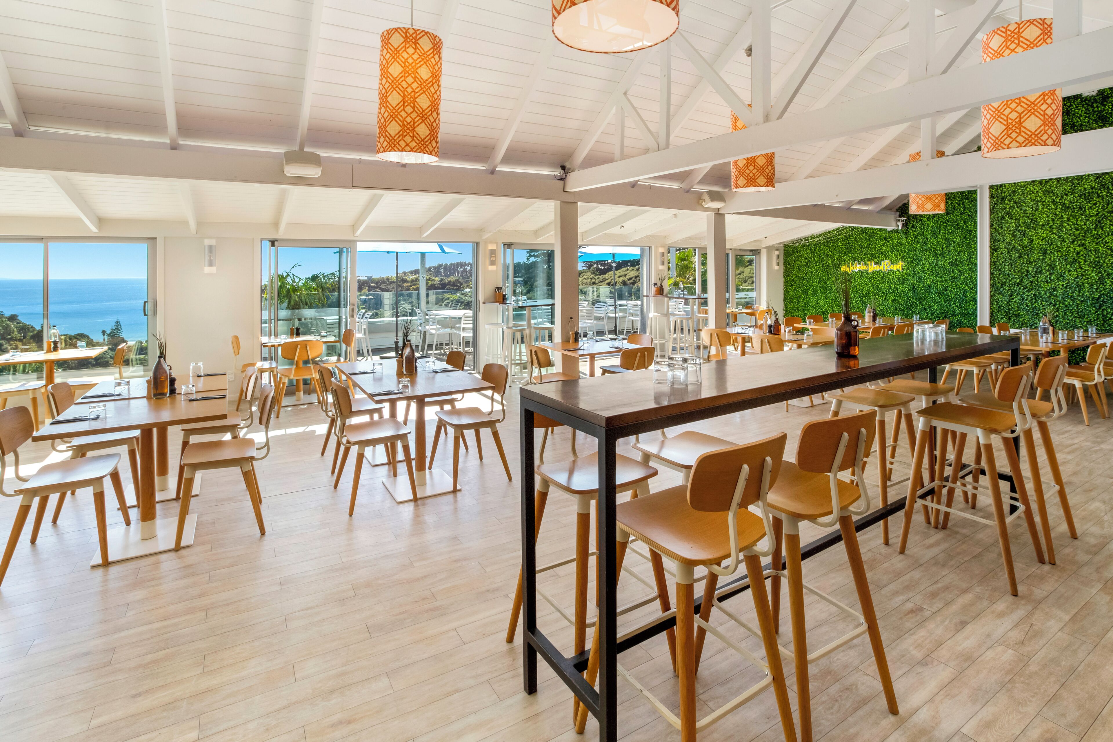A spacious dining area with high ceilings, large windows, and a view of the ocean. Tables and chairs are arranged for dining.