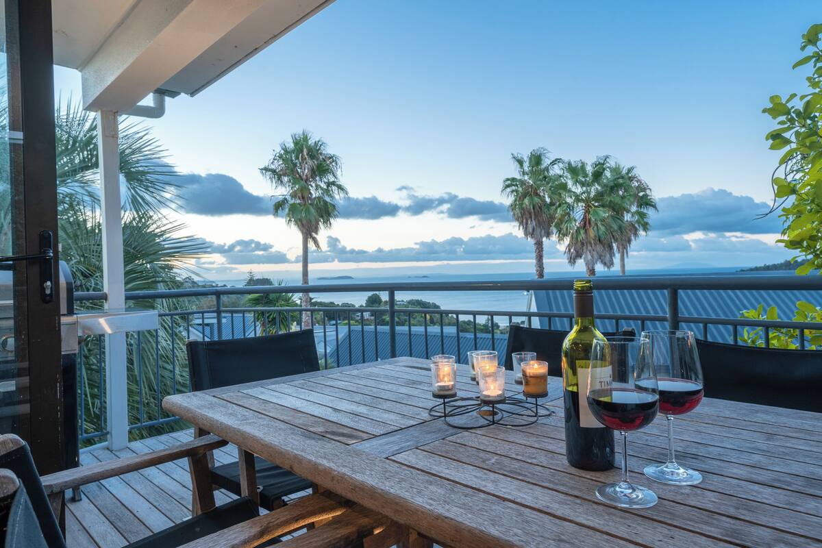 A wooden table with a bottle of wine and two glasses on a balcony overlooking the sea.