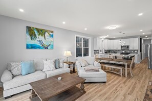 Ein modernes Wohnzimmer mit einer Couch, einem Hocker und einem Couchtisch. Im Hintergrund steht ein Esstisch mit Stühlen. Der Küchenbereich ist sichtbar, mit weißen Schränken und einer Mikrowelle.