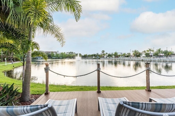 A waterfront deck with lounge chairs, a rope barrier, and a calm lake with a fountain.