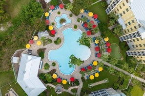 Vista aérea de un resort con una piscina central, varias zonas de descanso con sombrillas y abundante vegetación circundante.