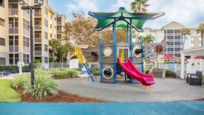 Un parque infantil con toboganes y estructuras para escalar, rodeado por una zona vallada y un edificio al fondo.