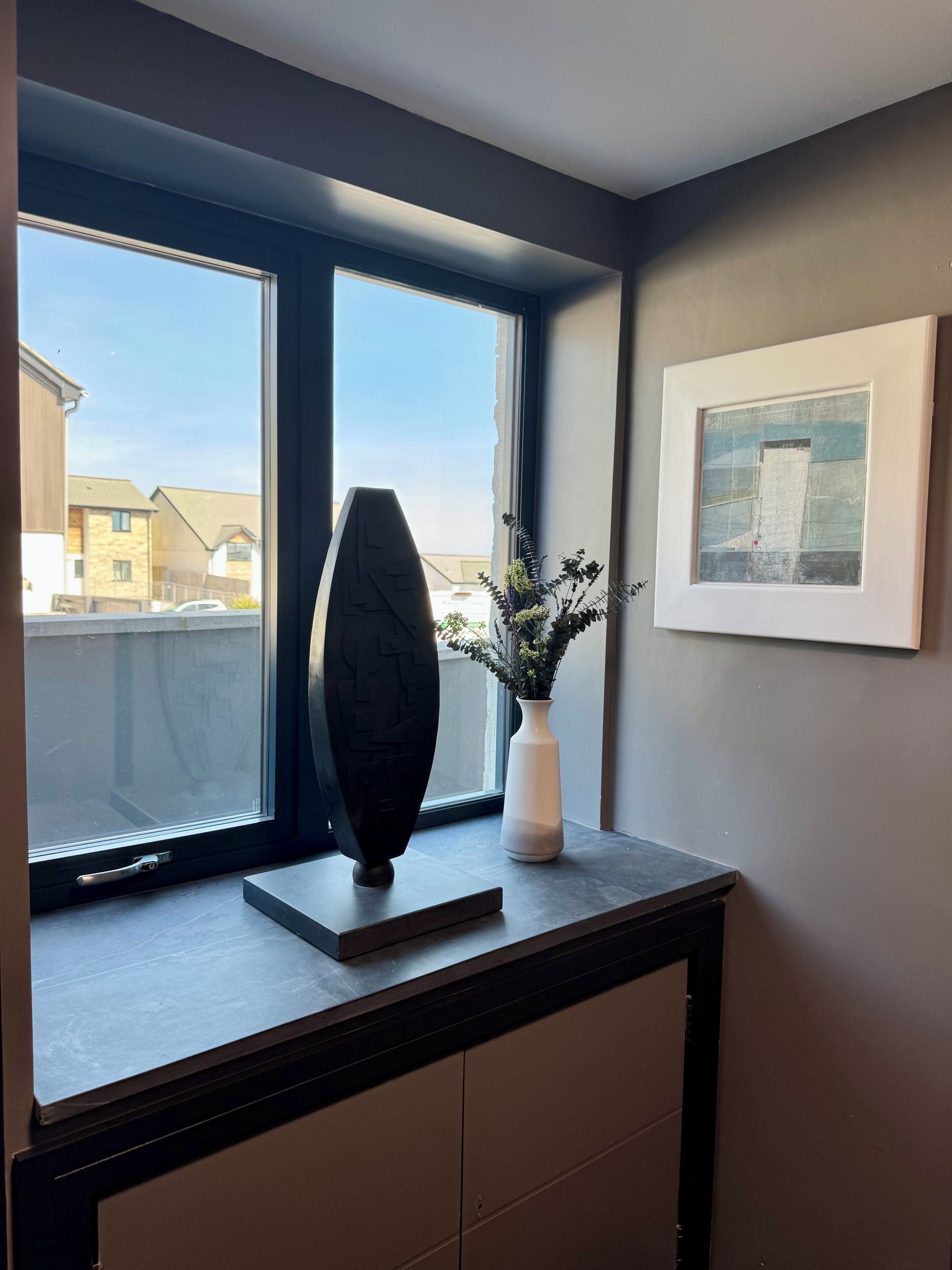 Une pièce avec une fenêtre, un vase rempli de fleurs et un tableau encadré accroché au mur.