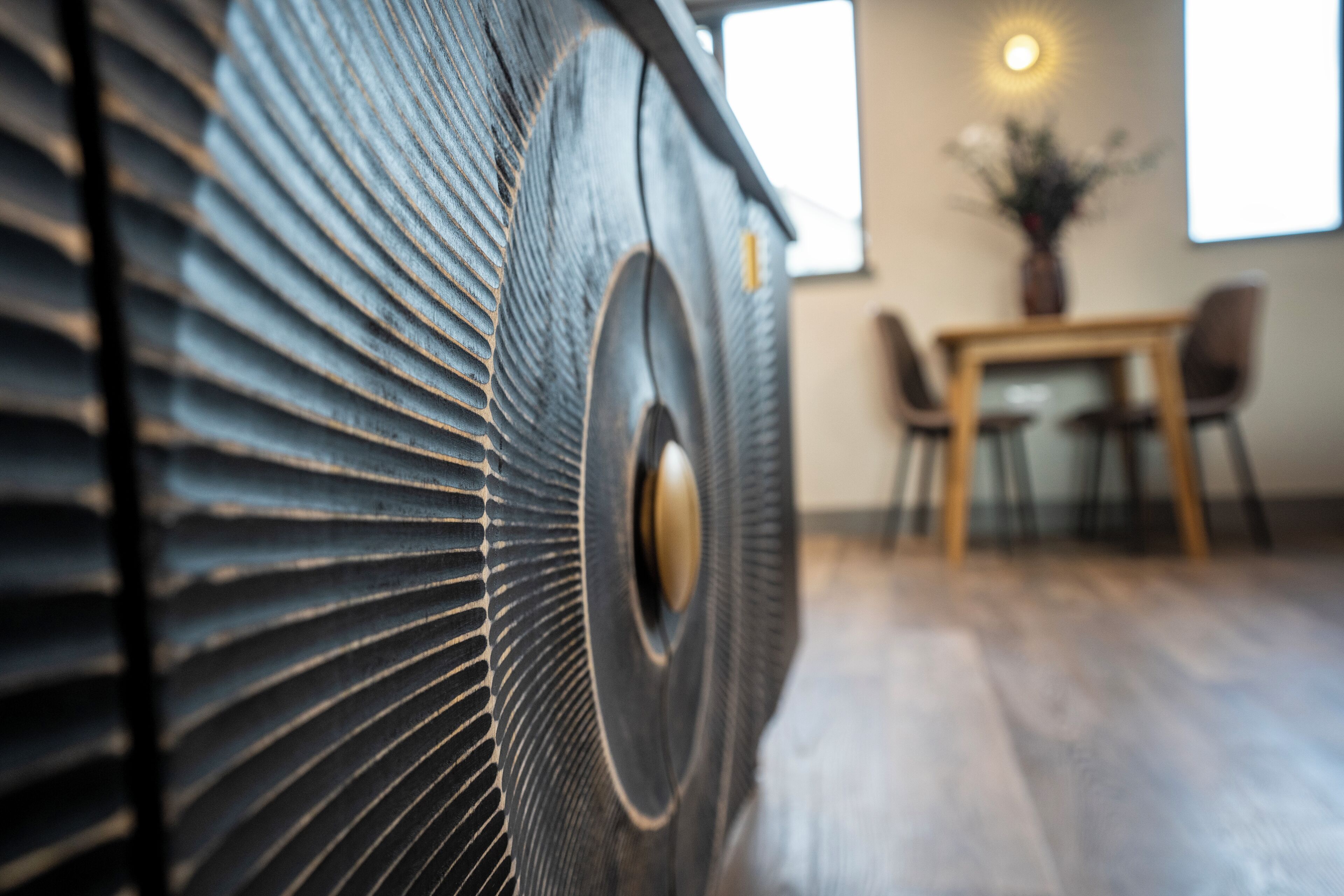 A close-up of a textured, metallic surface with a circular design element, likely part of a piece of furniture or decor. In the background, there is a dining area with a table and chairs, and a window allowing natural light to enter.