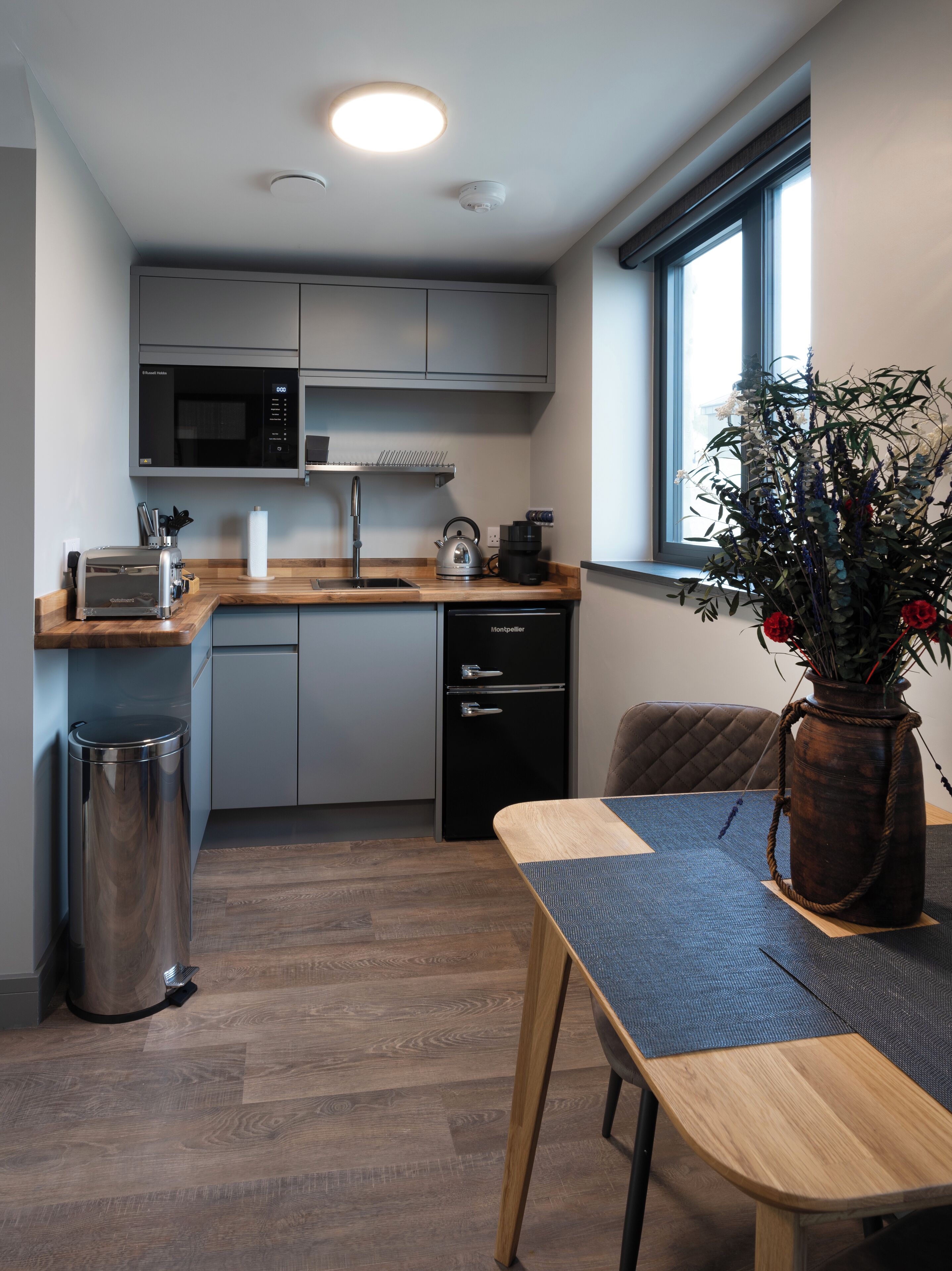 A compact kitchen with a microwave, toaster, sink, and dishwasher. There's a window with a view of greenery and a vase with flowers on the table.
