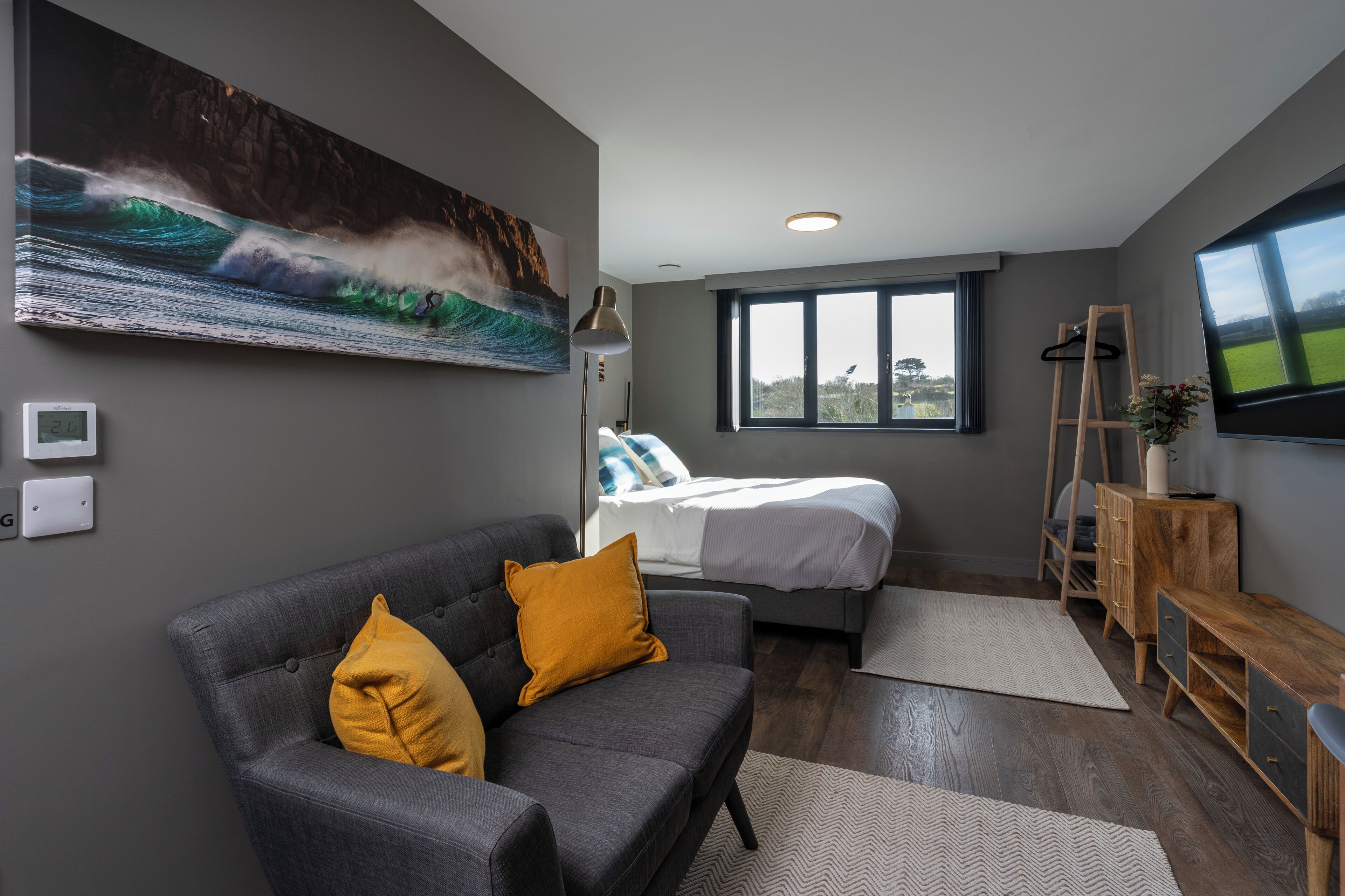Une chambre moderne avec un lit, un canapé gris agrémenté de coussins jaunes, une commode en bois, un téléviseur et une grande fenêtre.