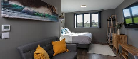 Um quarto moderno com uma cama, um sofá cinza com almofadas amarelas, um aparador de madeira, uma televisão e uma janela grande.