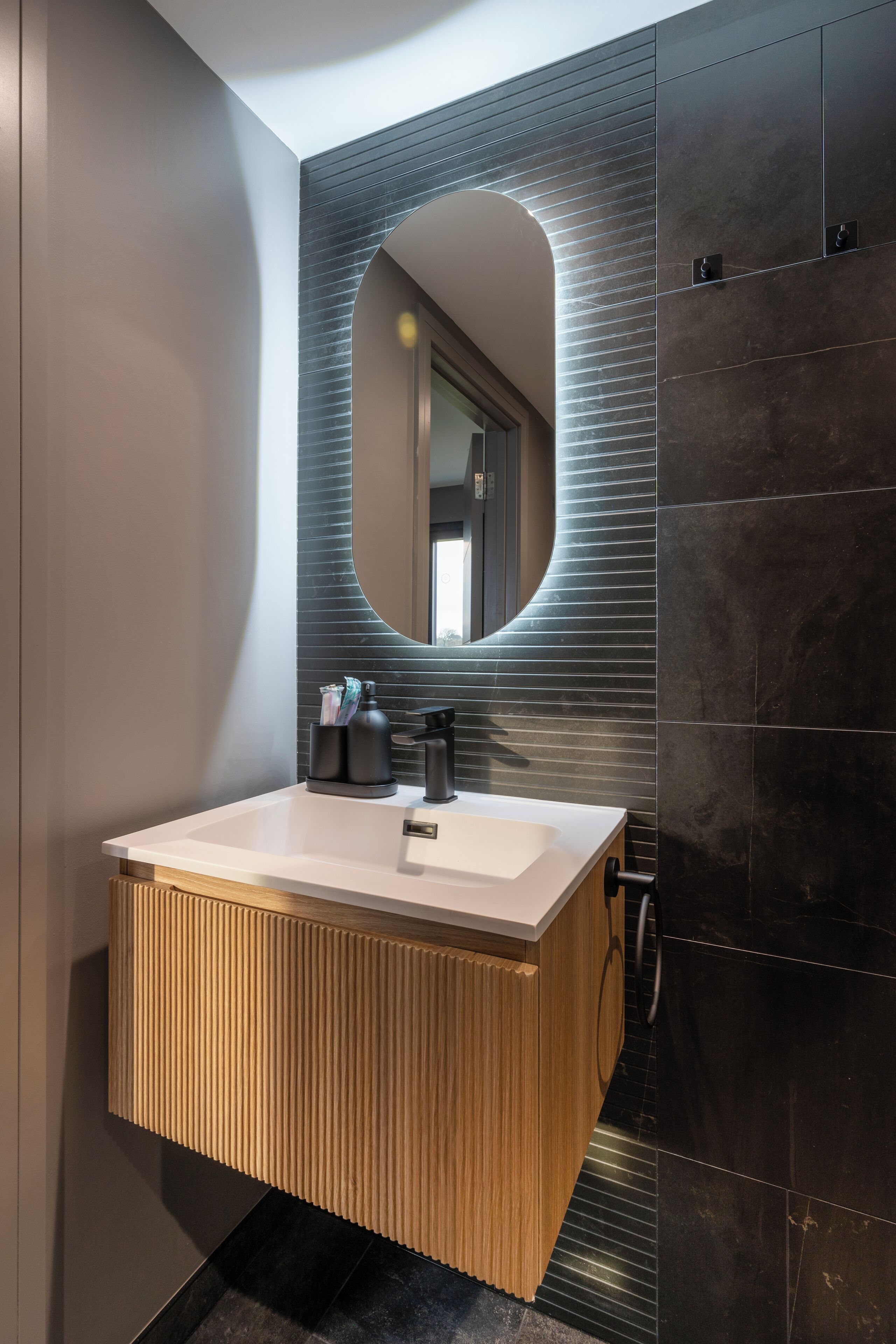 Un baño moderno con lavamanos blanco, paredes de baldosas oscuras y tocador de madera.