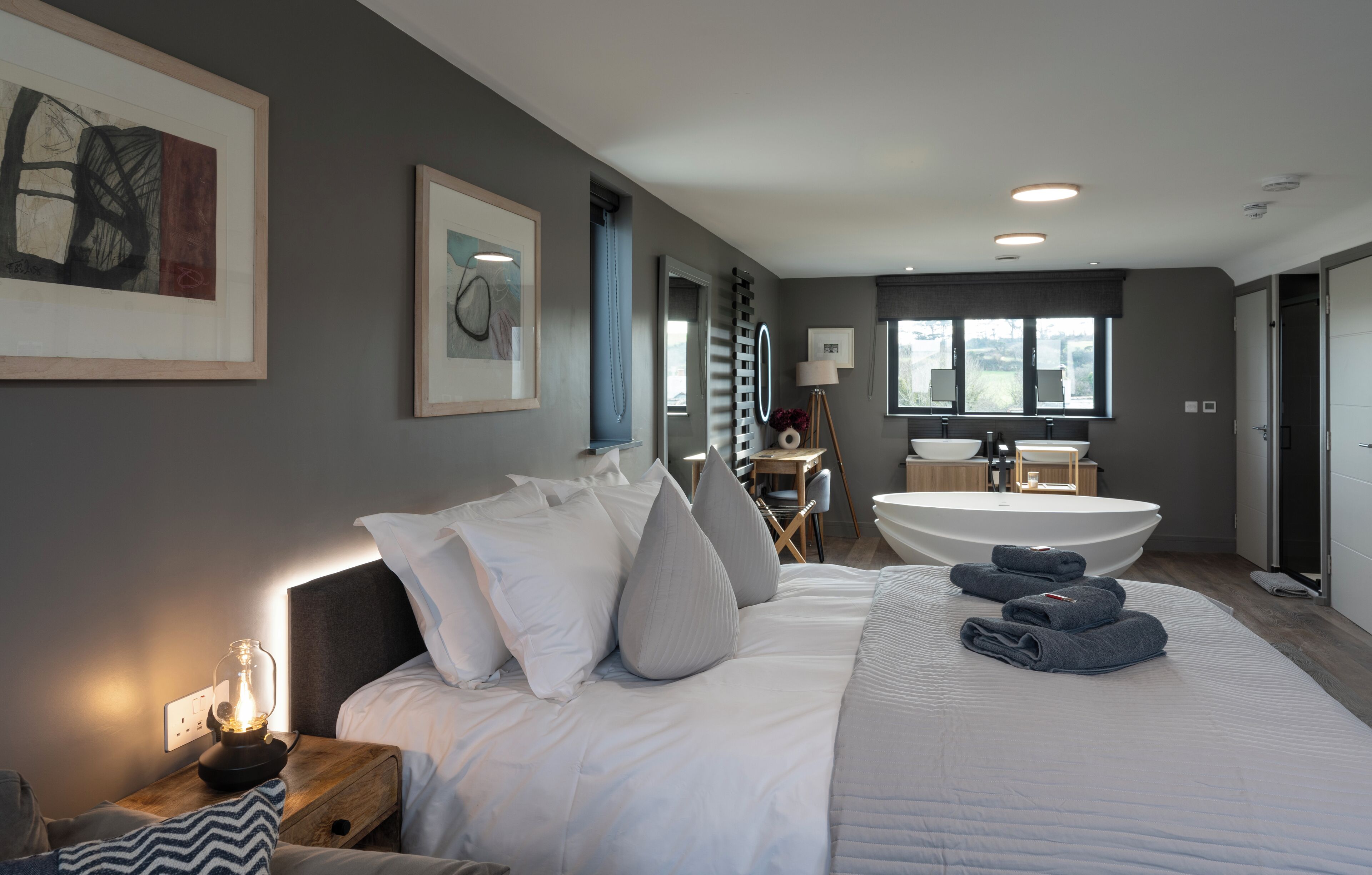 A modern bedroom with a large bed, a bedside table with a lamp, a bathroom with a bathtub, and artwork on the walls.