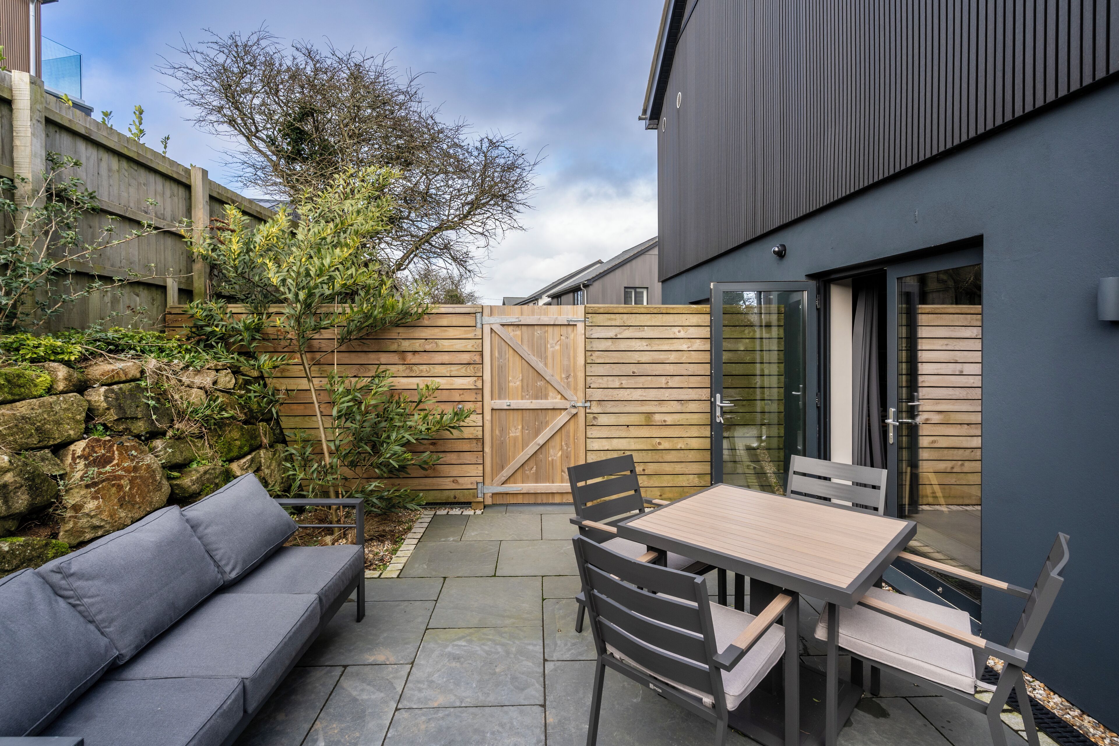 Une terrasse avec une table et des chaises en bois, un canapé gris et un mur en pierre.