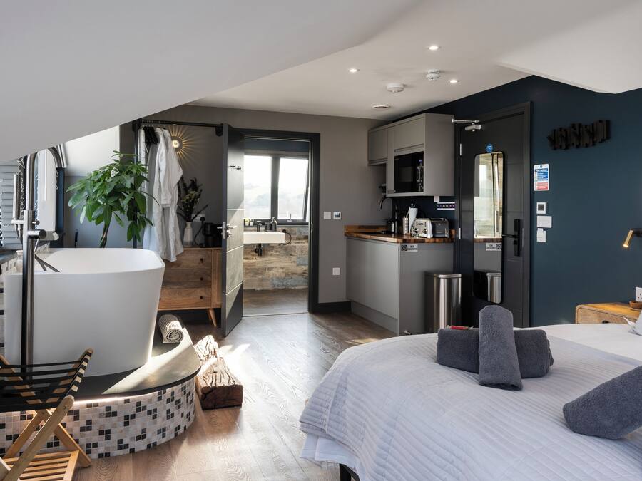 Una habitación de hotel moderna con una cama grande, una zona de estar con una silla, una pequeña cocina y un baño con bañera.
