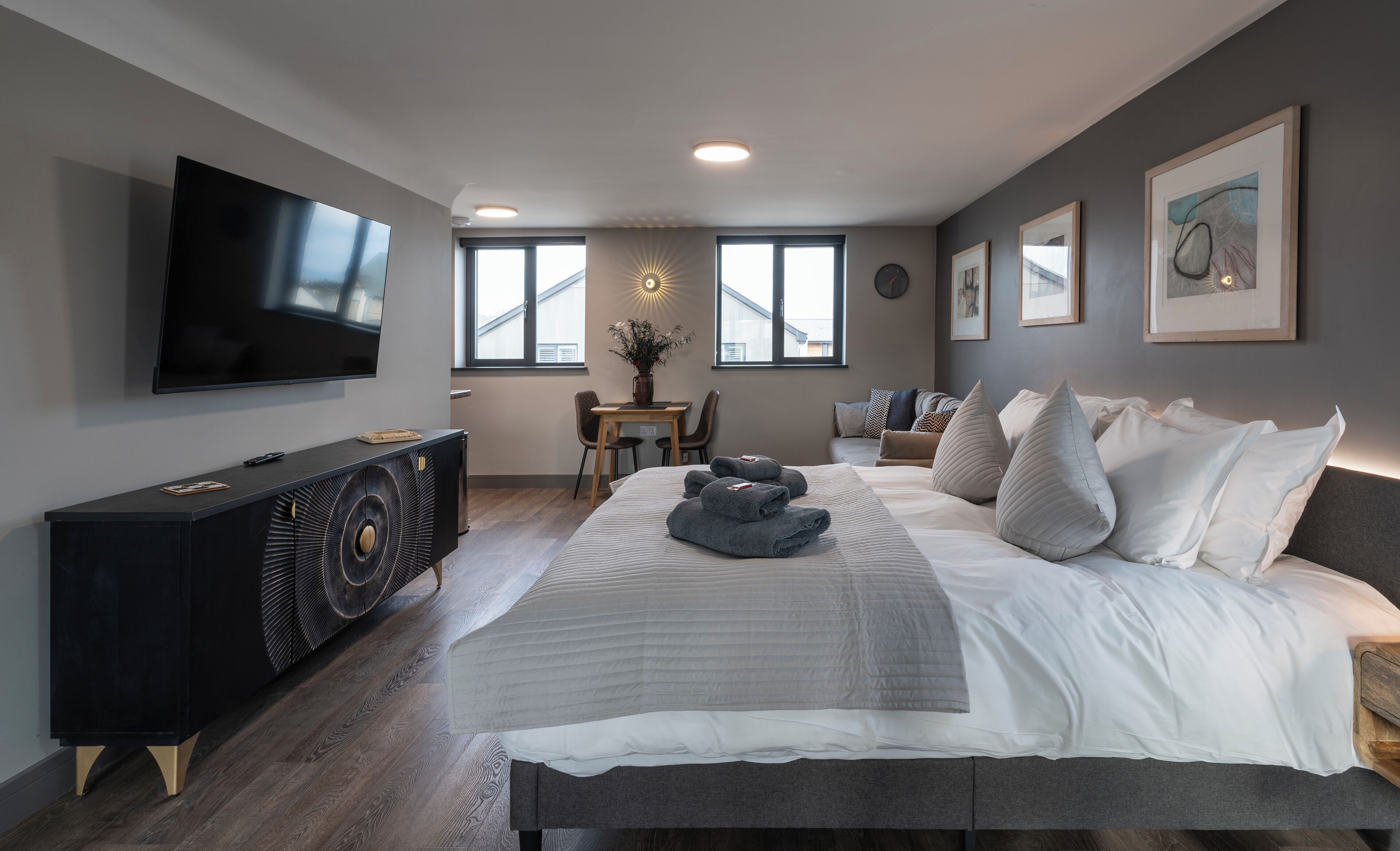 A modern bedroom with a large bed, a TV mounted on the wall, a dining area with a table and chairs, and a window with a view of buildings.