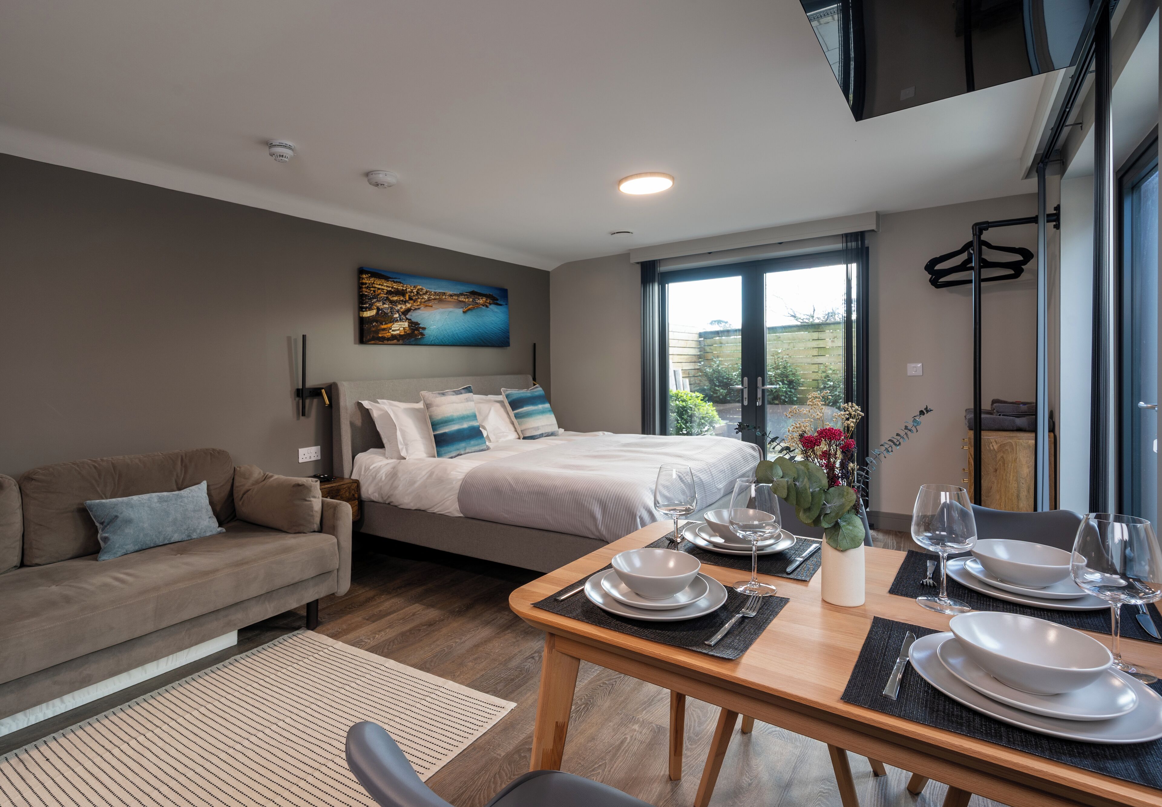 A modern hotel room with a bed, a sofa, a dining table set with plates and glasses, and a painting on the wall.