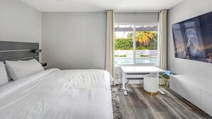 Moderni hotellihuone, jossa on suuri sänky, työpöytä ja näkymä uima-altaalle ja palmuille.