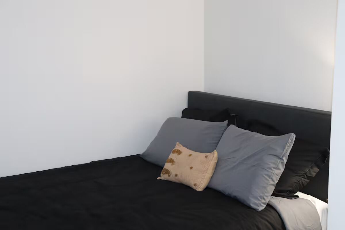 Una cama con ropa de cama negra y dos almohadas, una color beige y otra gris, en una habitación con paredes blancas.