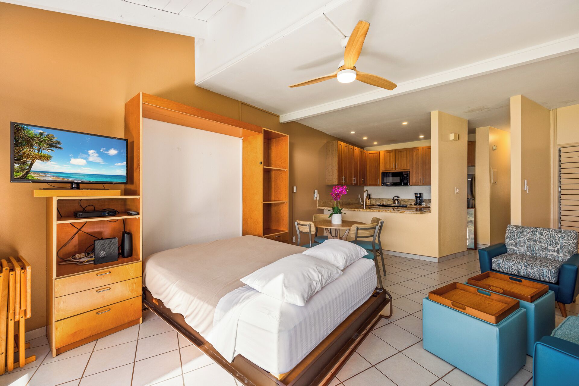 Une chambre avec un lit, une télévision, un bureau et une chaise. Il y a également un coin cuisine avec une table et des chaises.