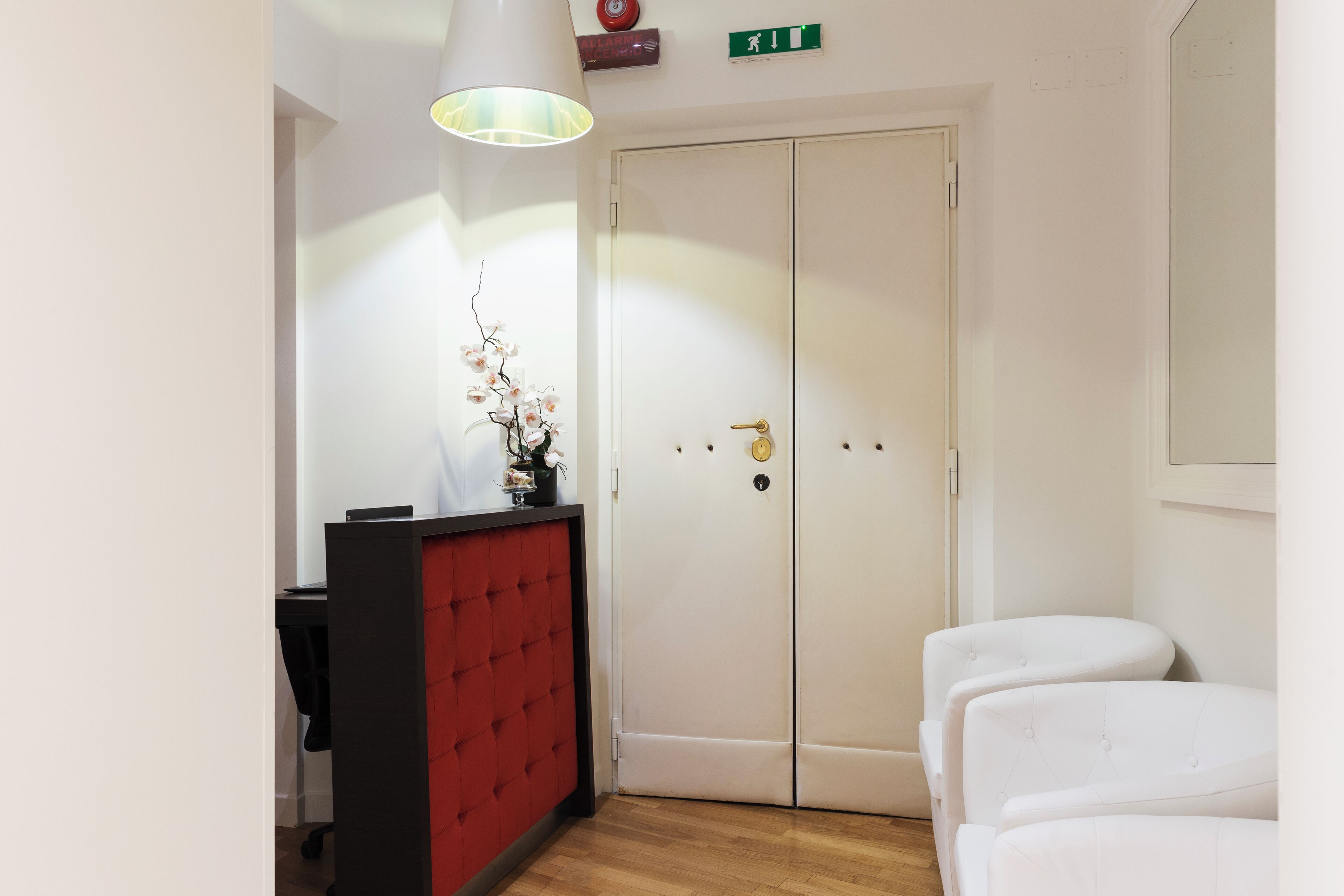 A hotel room with a red and black desk, a white sofa, a white wardrobe, and a hanging lamp.