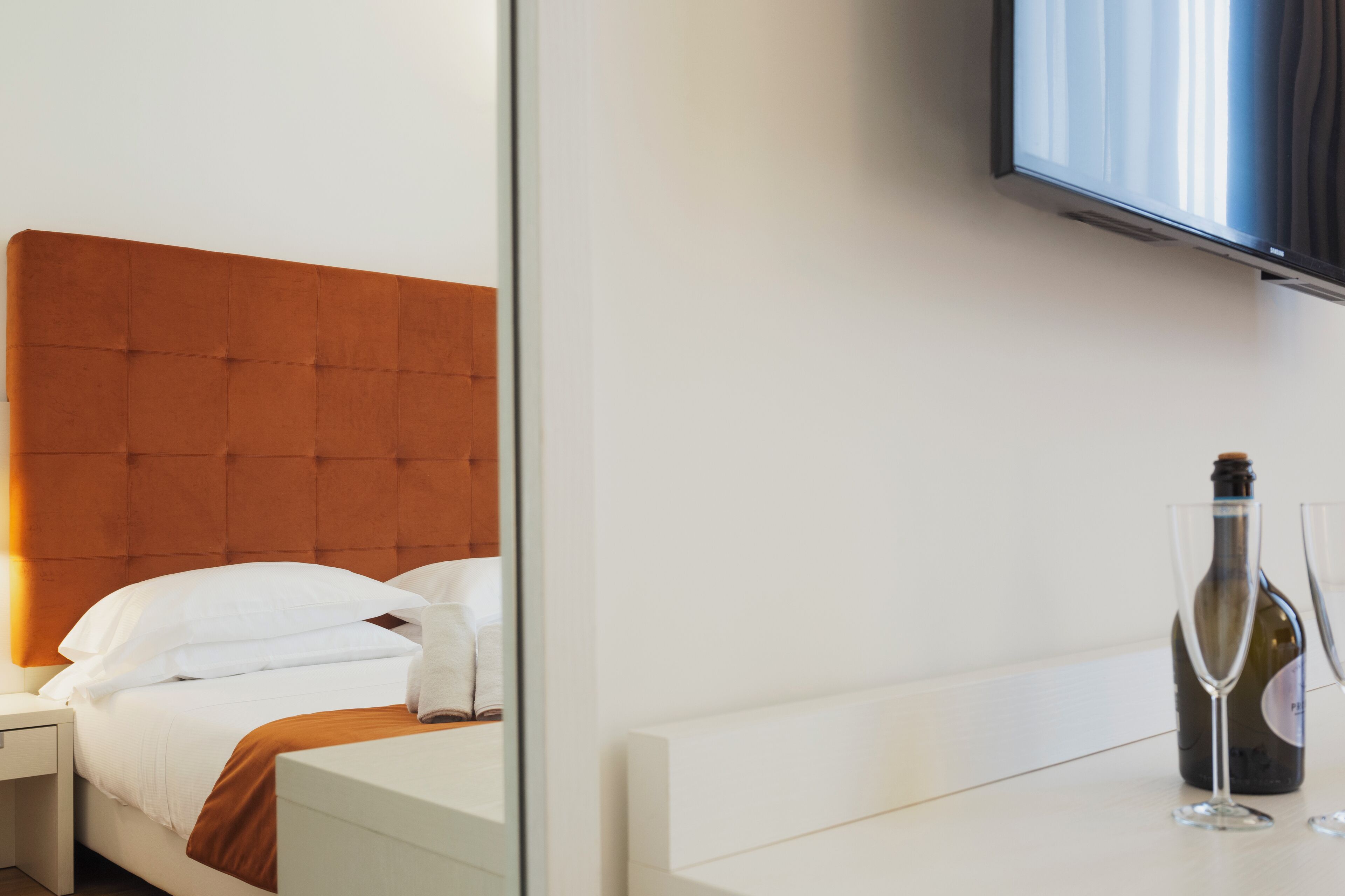 A modern hotel room with a large bed, a bedside table, a television mounted on the wall, and a bottle of wine on the table.