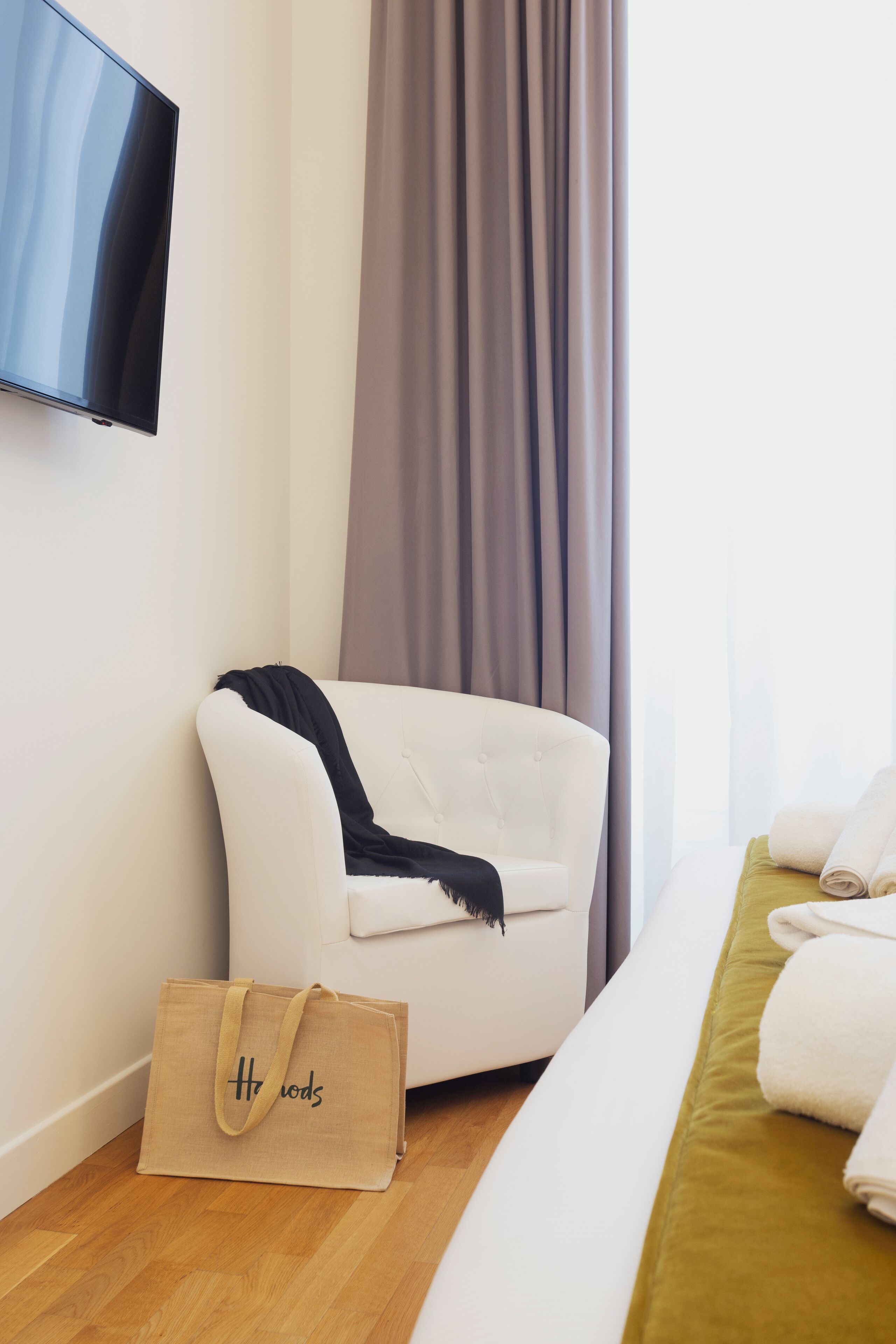 A hotel room with a white armchair, a bed with a green bedspread, a TV mounted on the wall, and a beige bag on the floor.