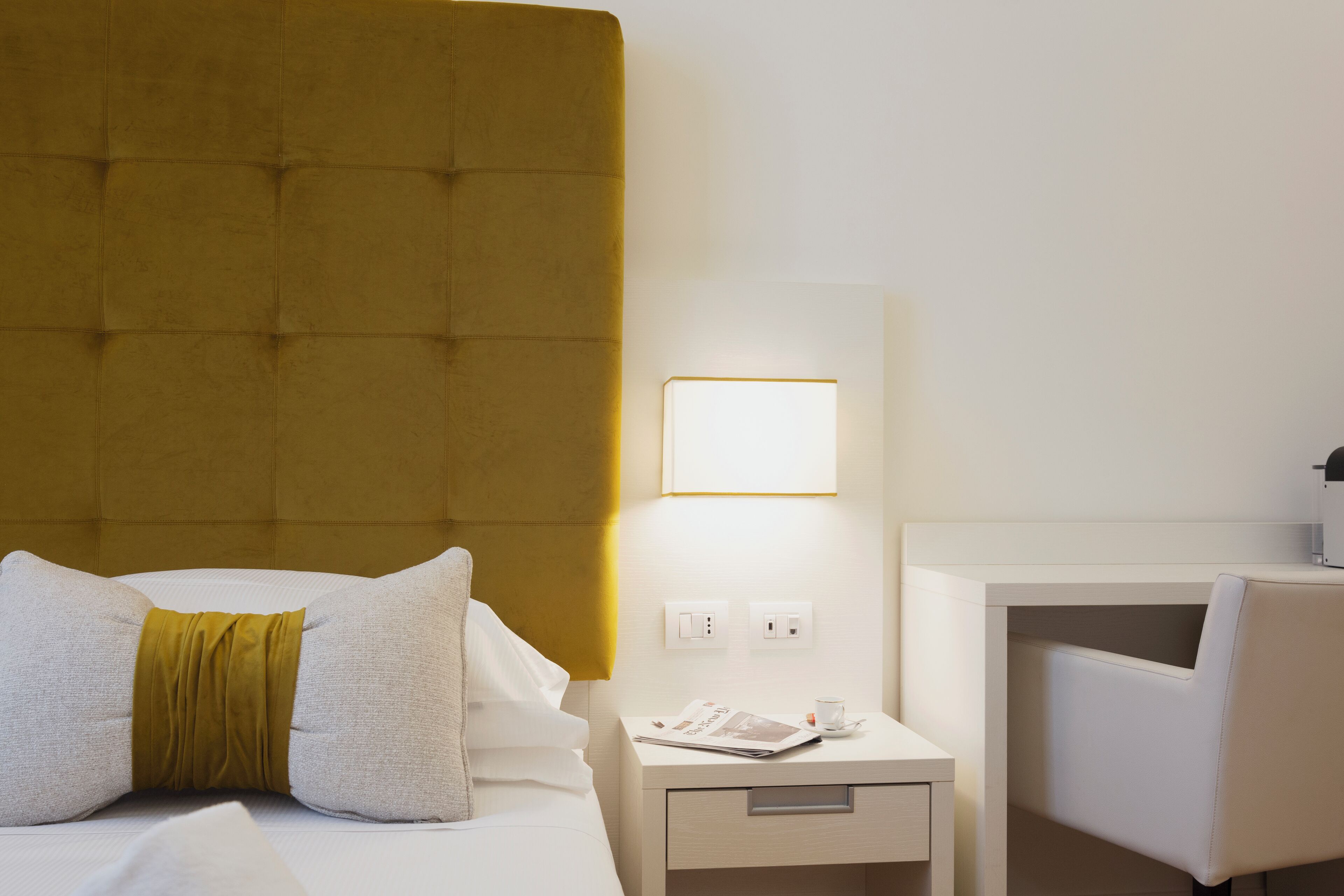 A hotel room with a tufted headboard, a bedside table with a lamp, a chair, and a nightstand with a phone and a cup.
