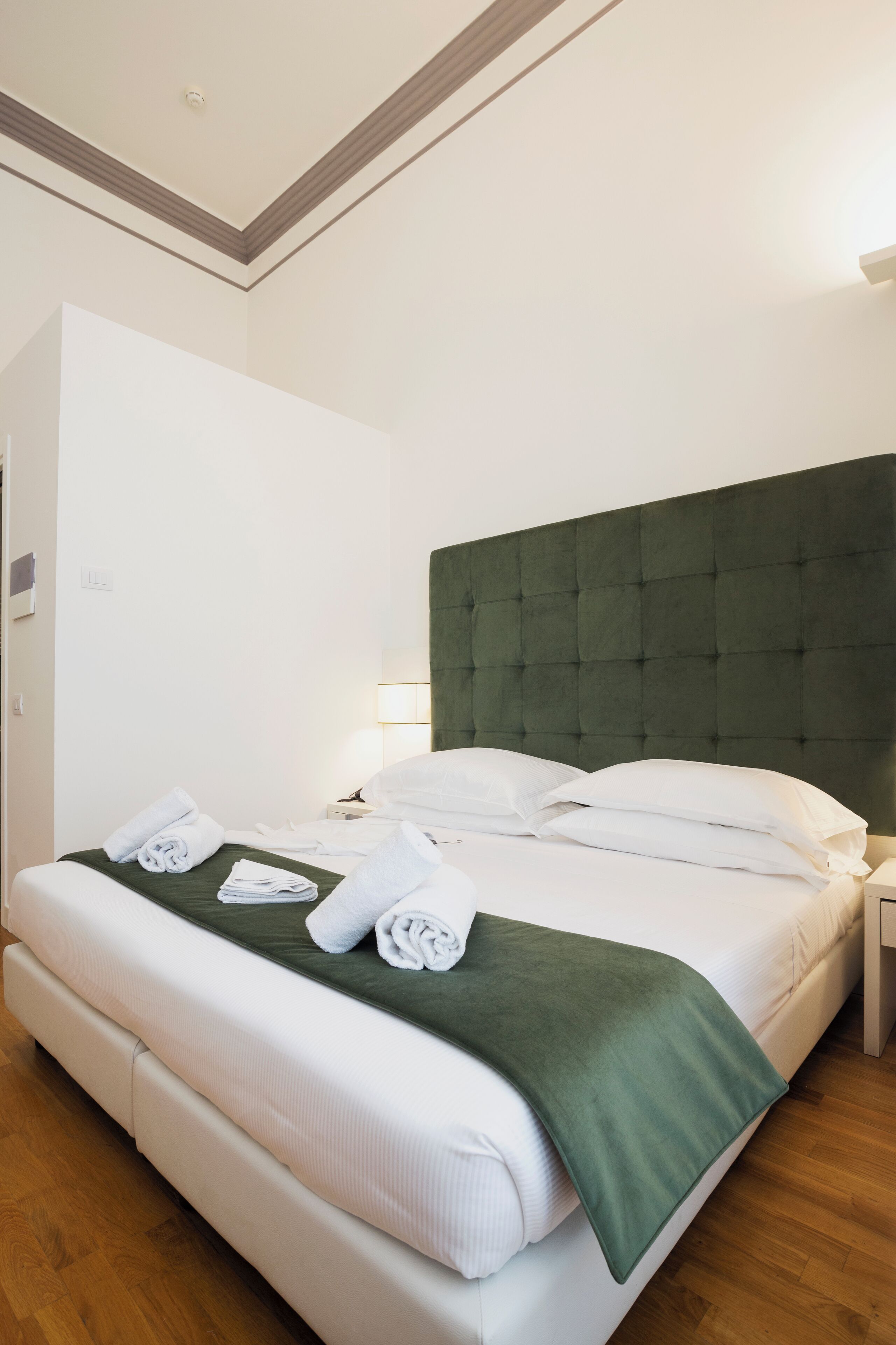 Una cama bien tendida con sábanas blancas y una manta verde, suelo de madera y pared blanca con una lámpara en el techo.