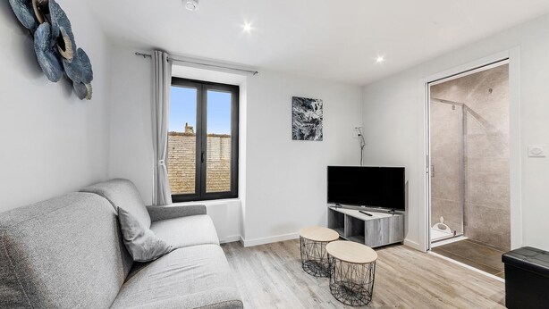 Una sala moderna con un sofá gris, un televisor de pantalla plana, dos mesitas redondas y un muro cortinero que da a una zona de ducha.