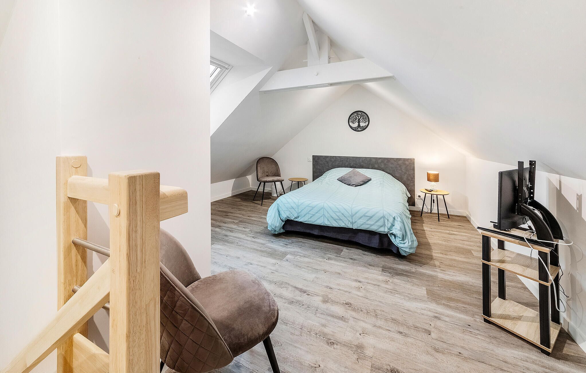 A bedroom with a sloped ceiling, a bed with a blue bedspread, a chair, a small table, and a TV stand.