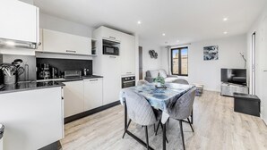 Una cocina moderna con armarios blancos, horno integrado y microondas. Hay un comedor con mesa y sillas, y un televisor en una esquina.