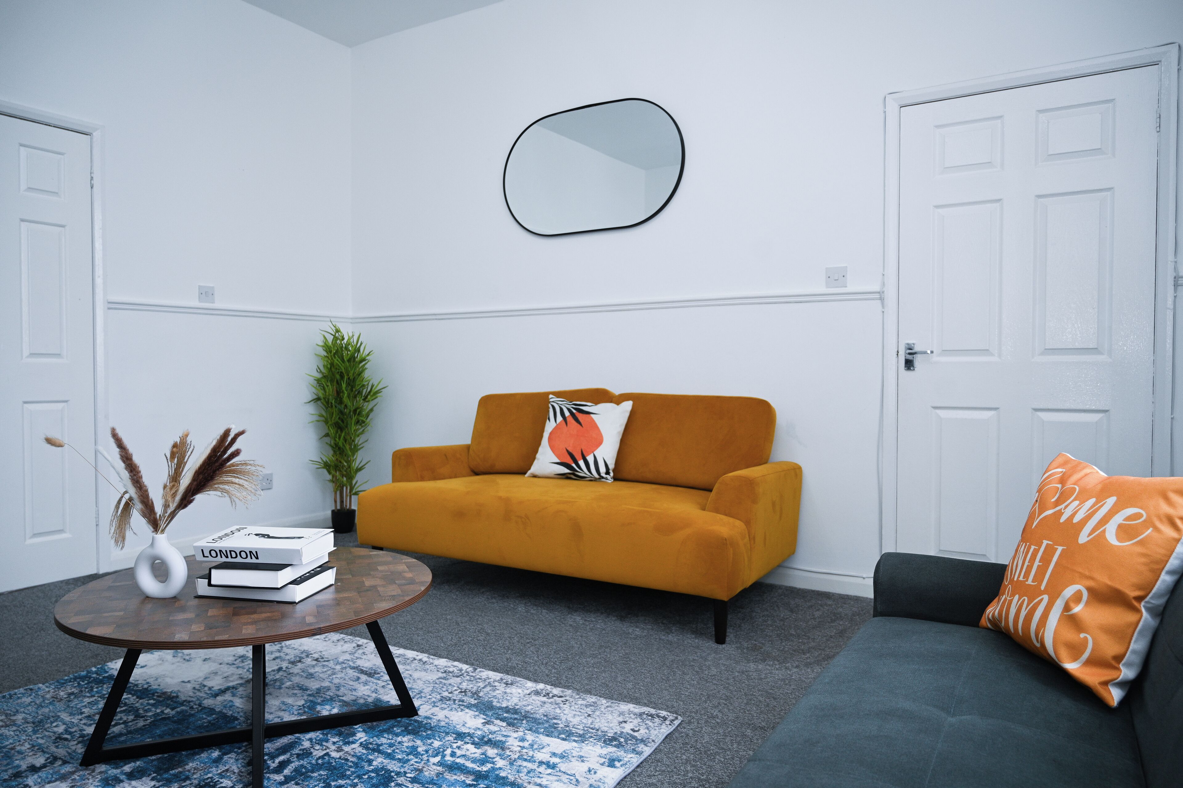Een woonkamer met een mosterdgele bank, een rond bijzettafeltje en een blauw vloerkleed.