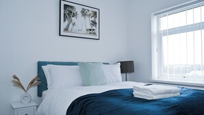 Una cama bien hecha con un cabecero azul, un cuadro enmarcado con una playa y una ventana con persianas.
