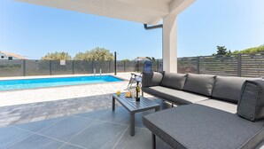 A covered outdoor area with a swimming pool, a sofa, and a table with a bottle and glass.