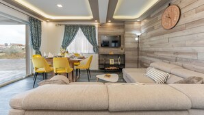A modern living room with a sofa, dining table, and chairs. A TV is mounted on the wall, and there is a fireplace.