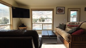 Ein Wohnzimmer mit einer Ledersofa, einem Holztisch, einem flachbildfernseher an der Wand und einem großen Fenster mit Blick auf eine gebirgige Landschaft.