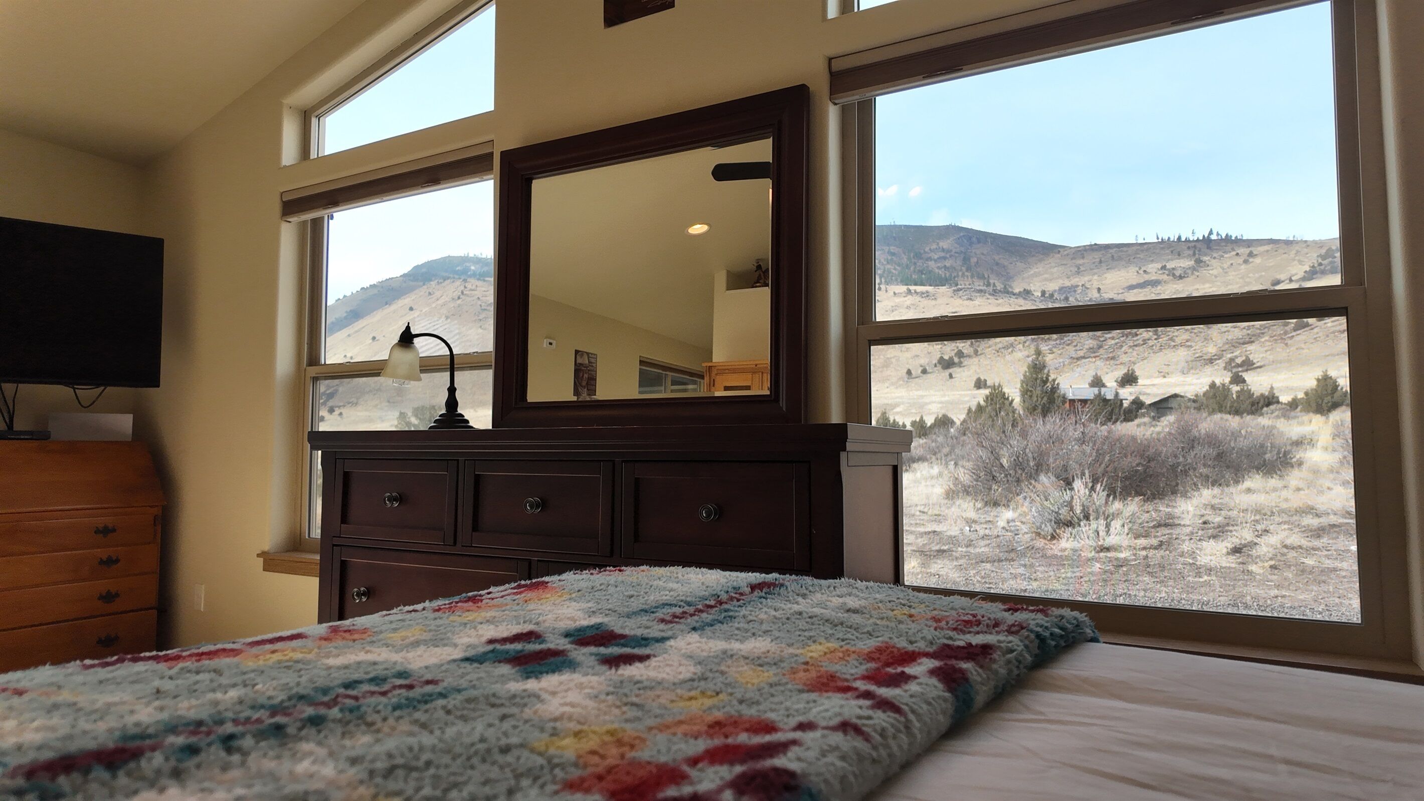 A bedroom with a large window, a dresser, a bed with a colorful blanket, and a flat-screen TV on a stand.