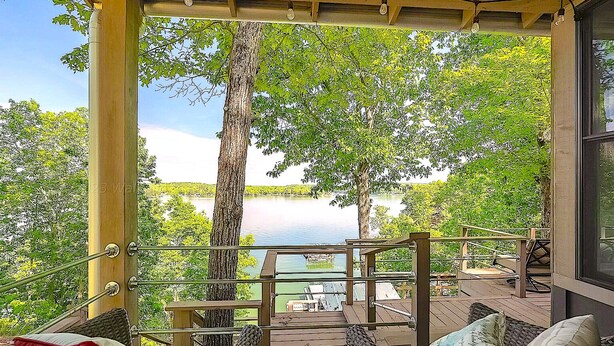 Un espacio de estar techado al aire libre con mobiliario de mimbre, con vistas a un lago y árboles.