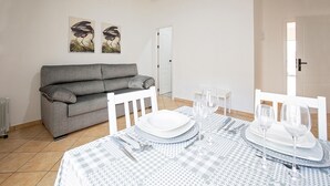 Ein Essbereich mit kariertem Tischleinen, weißen Stühlen und einem grauen Sofa. An der Wand hängen zwei gerahmte Vogelbilder.