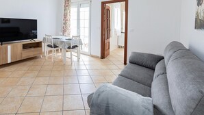 Ein Wohnzimmer mit grauem Sofa, einem Fernseher auf einem Holzschrank, einem Esstisch mit Stühlen und einer Tür zu einem weiteren Raum.