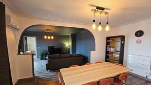 A modern living room with a wooden dining table, orange chairs, and a black sofa. There is a bookshelf and a TV in the background. - 3-bedroom 110m² apartment with terrace, view (Thiers)
