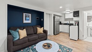A modern living room with a dark blue accent wall, a brown sofa with cushions, a round coffee table, and a kitchen area with white cabinets and appliances. - Cozy 1 Bed, 1 Bath - Near Downtown and U of U. (Salt Lake City)