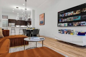 A modern living room with a sofa, dining area, kitchen, and a wall-mounted TV displaying various shows.
