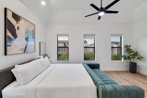 A modern bedroom with a large bed, a ceiling fan, and three windows offering views of buildings and greenery. - Downtown~3 Bedroom Ensuite~Walkable~Rooftop! (Dallas)