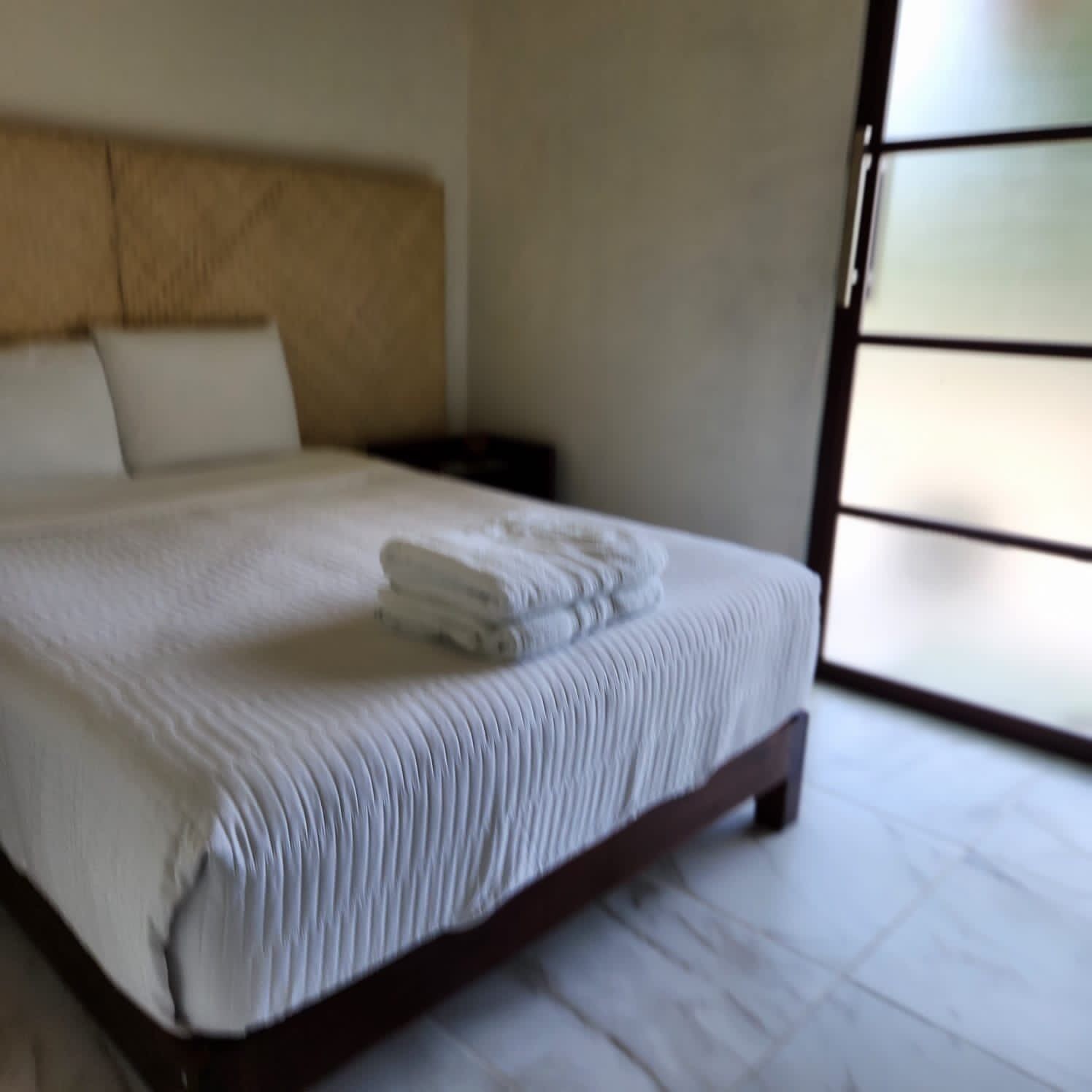 Uma cama bem arrumada com edredom branco e toalhas dobradas, cabeceira de madeira e mesa de cabeceira com abajur.