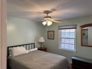 Ein Schlafzimmer mit Bett, Nachttisch, Lampe, Deckenventilator, Fenster mit Aussicht und Kommode.