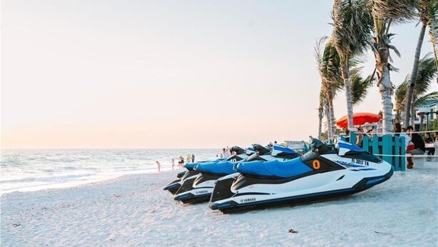 Jet skis lined up on a sandy beach with palm trees in the background.