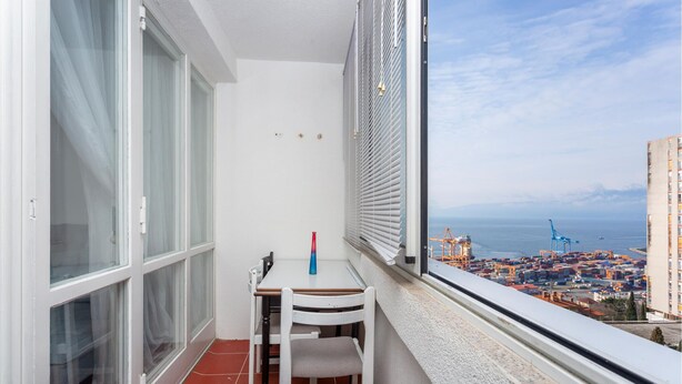 Een balkon met een tafel en stoelen, een uitzicht op een haven met containers en kranen, en een gebouw op de achtergrond.